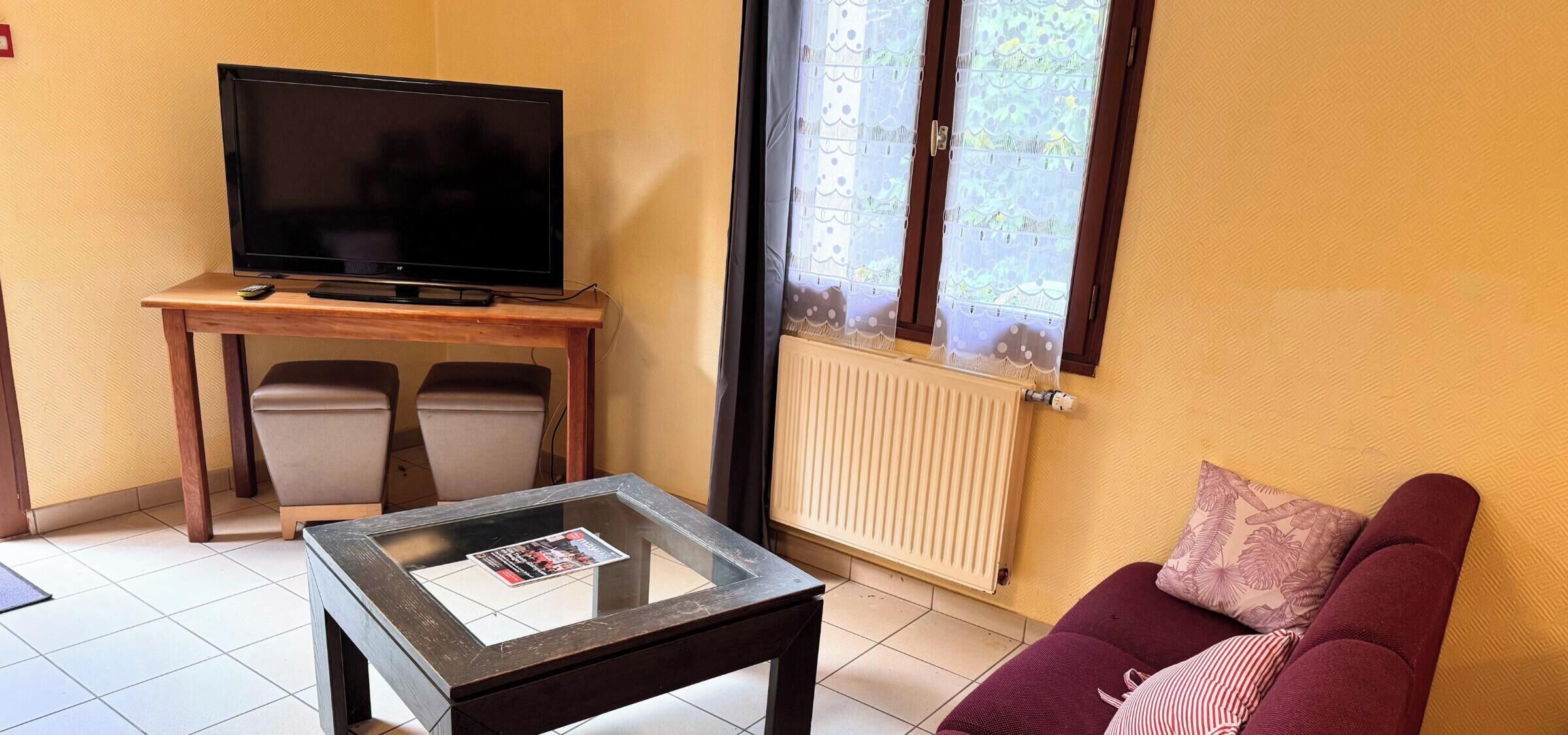 Een woonkamer met een televisie op een houten tafel, een glazen bijzettafeltje, een bank met een kussen en een raam met uitzicht op groen.