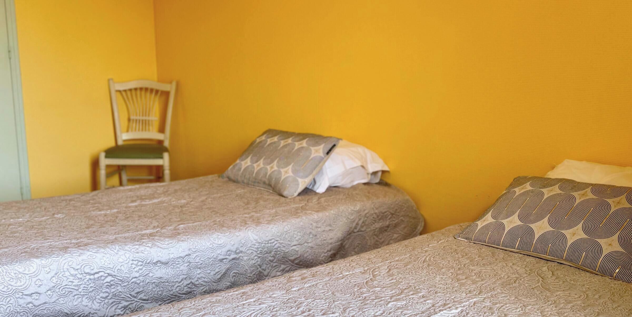 A room with two beds, each with patterned bedding, a chair, and a yellow wall.