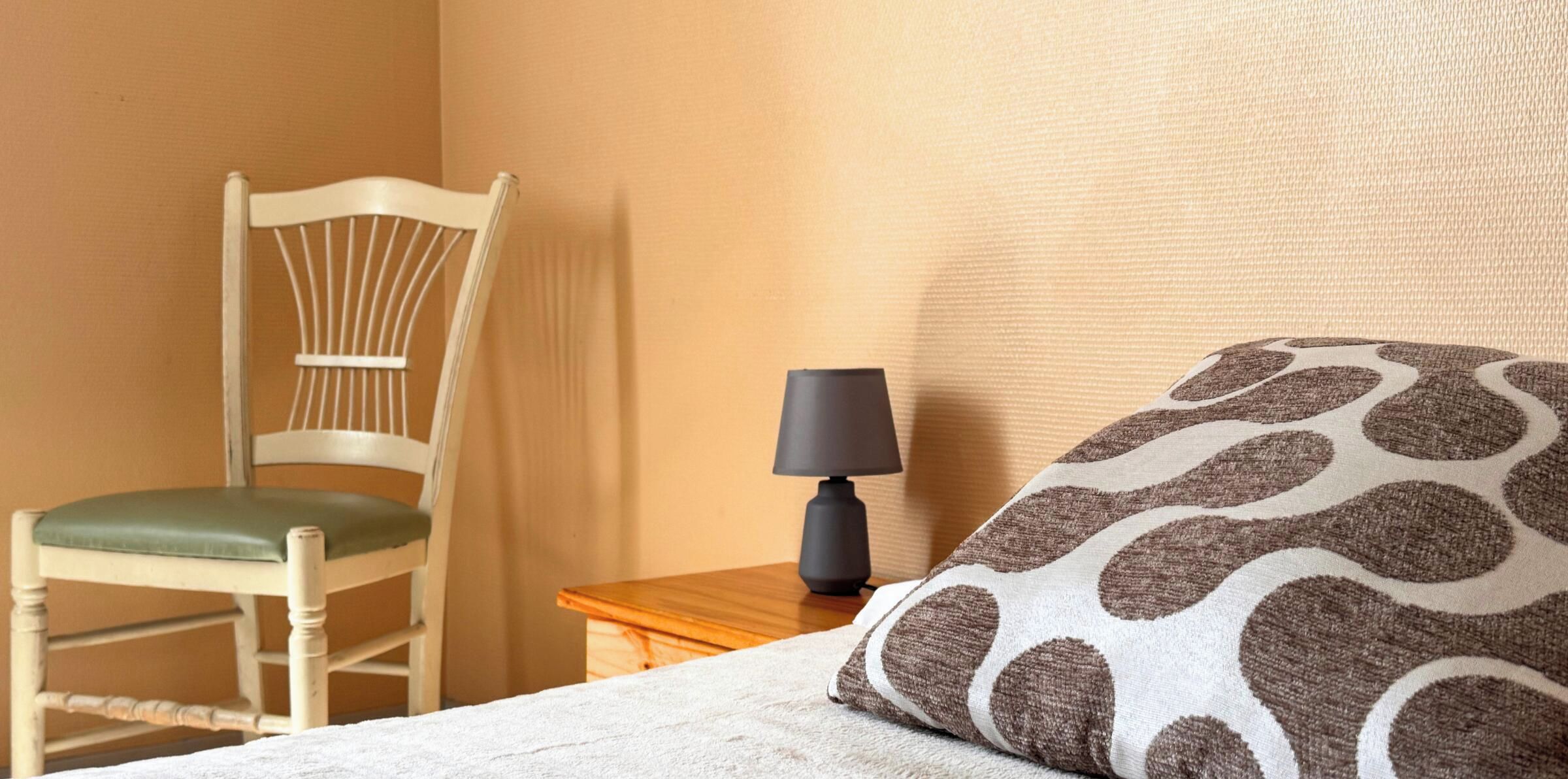 A hotel room with a wooden chair, a bedside table with a lamp, and a patterned pillow.