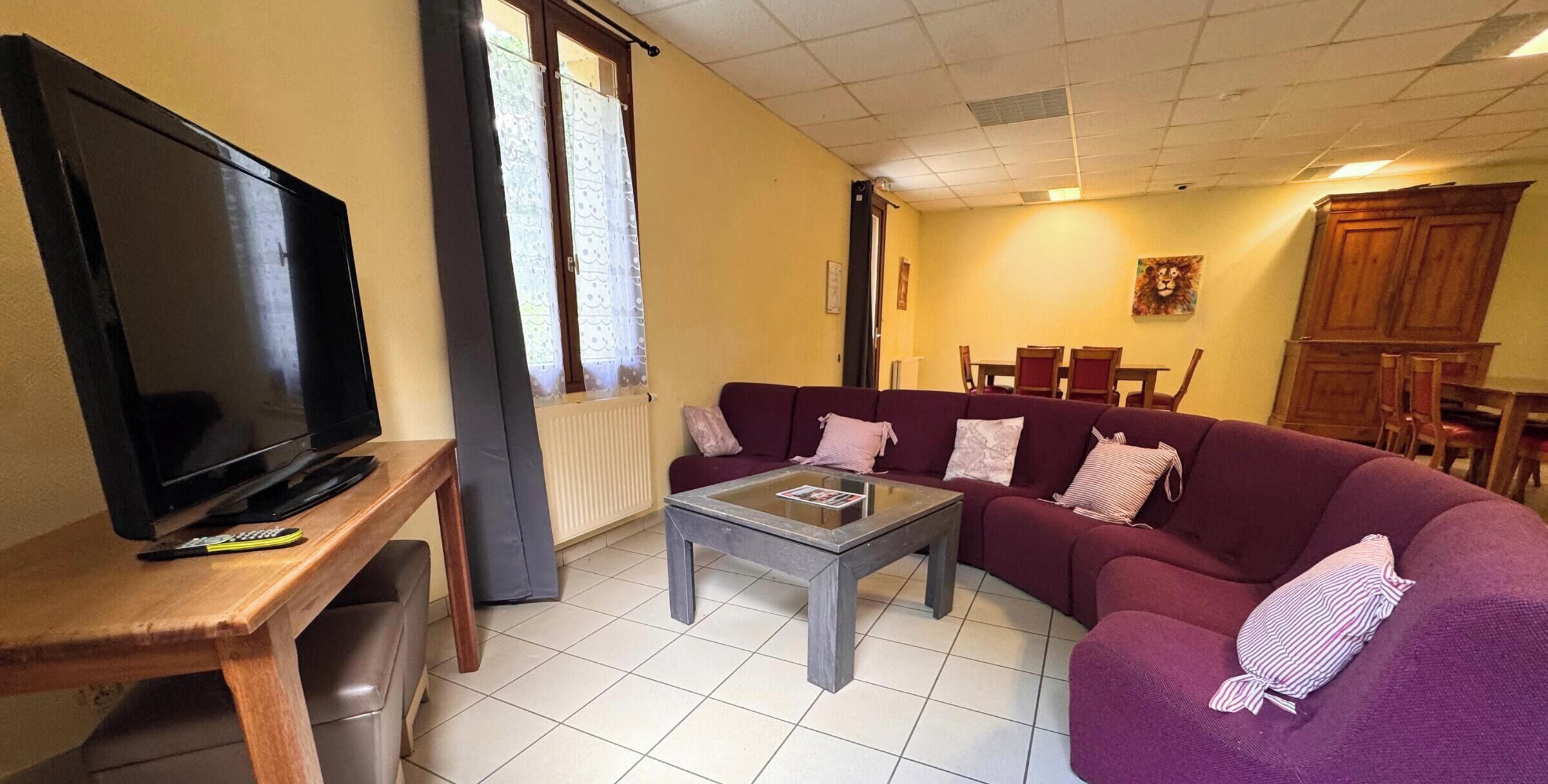 Un salon avec un téléviseur posé sur un meuble en bois, un canapé violet, une table basse en verre et, au fond, un coin repas.
