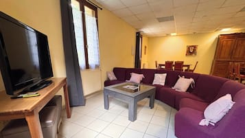 Un soggiorno con televisione su un mobile in legno, un divano viola, un tavolino da caffè in vetro e un'area pranzo sullo sfondo.