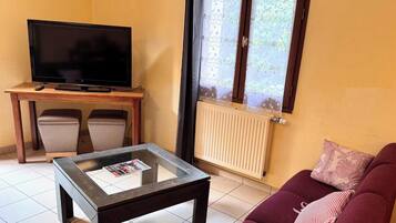 Un soggiorno con televisione su un tavolino di legno, un tavolino da caffè in vetro, un divano con un cuscino e una finestra vista verde.