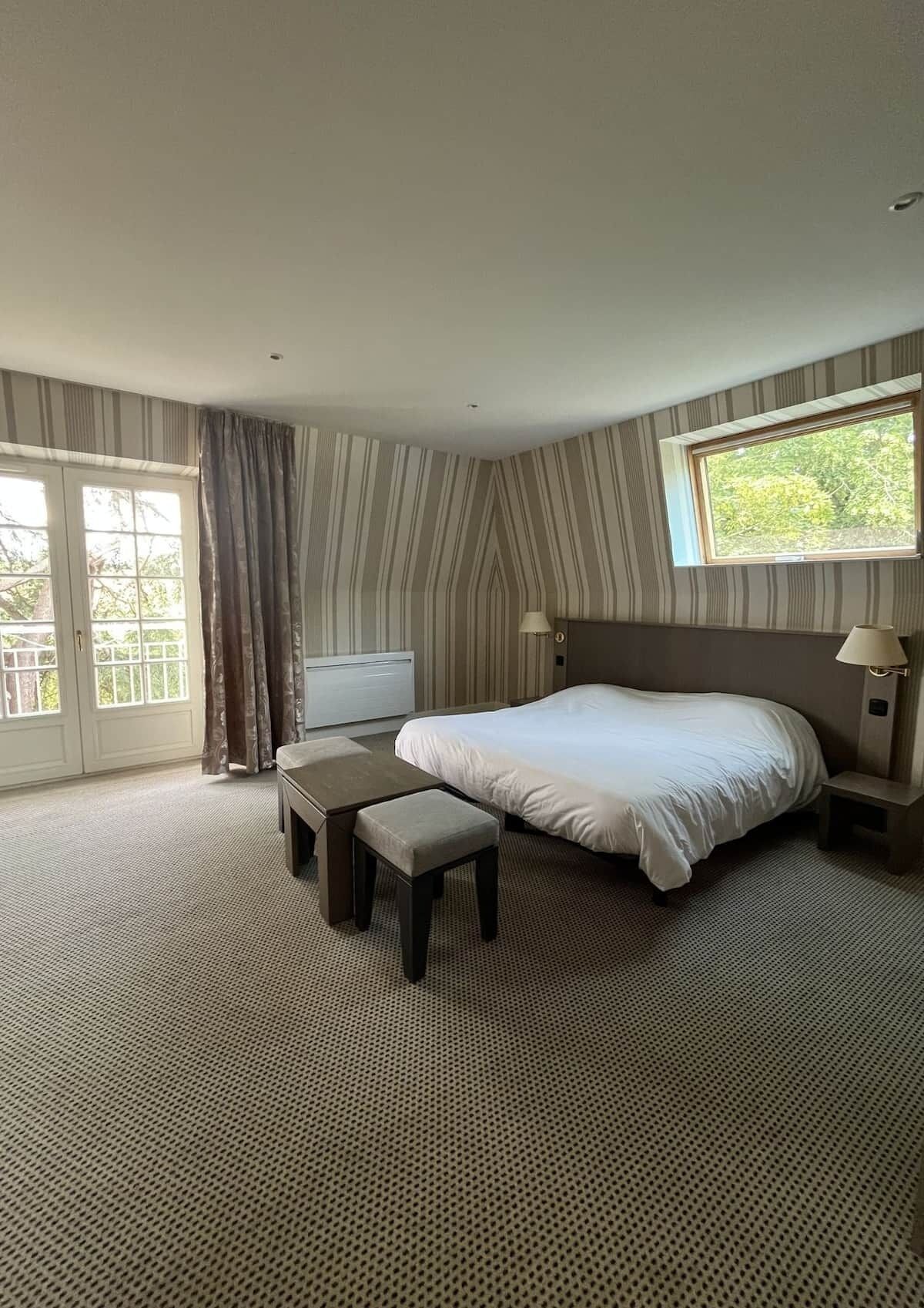 A hotel room with a large bed, a desk, a chair, a TV, and a window with a view of trees.