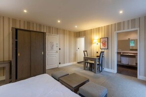 A hotel room with a bed, a desk, a chair, a small table, a bench, and a bathroom with a mirror and sink.