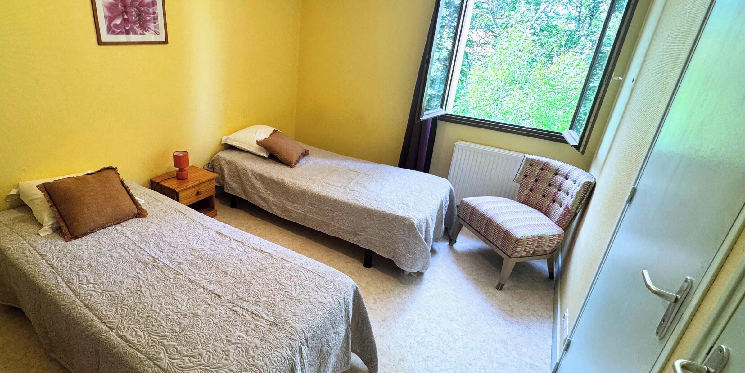 A small, cozy room with two single beds, a nightstand, a chair, and a window with a view of greenery.