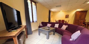 Ein Wohnzimmer mit Fernseher auf einem Holzständer, einem violetten Sofa, einem Glastisch und einem Essbereich im Hintergrund.