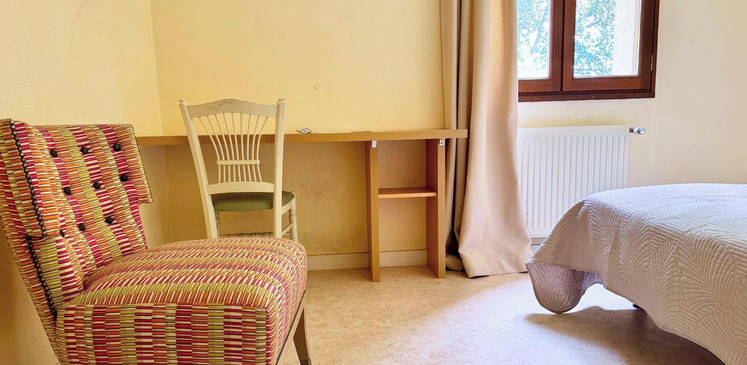 A room with a wooden desk, a chair with a striped cushion, a bed with a quilt, and a window with curtains.