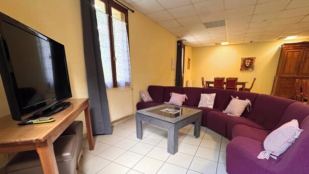 A living room with a television on a wooden stand, a purple sofa, a glass coffee table, and a dining area in the background. - Room 104 - Le Bas Mont Residence (Moulay)