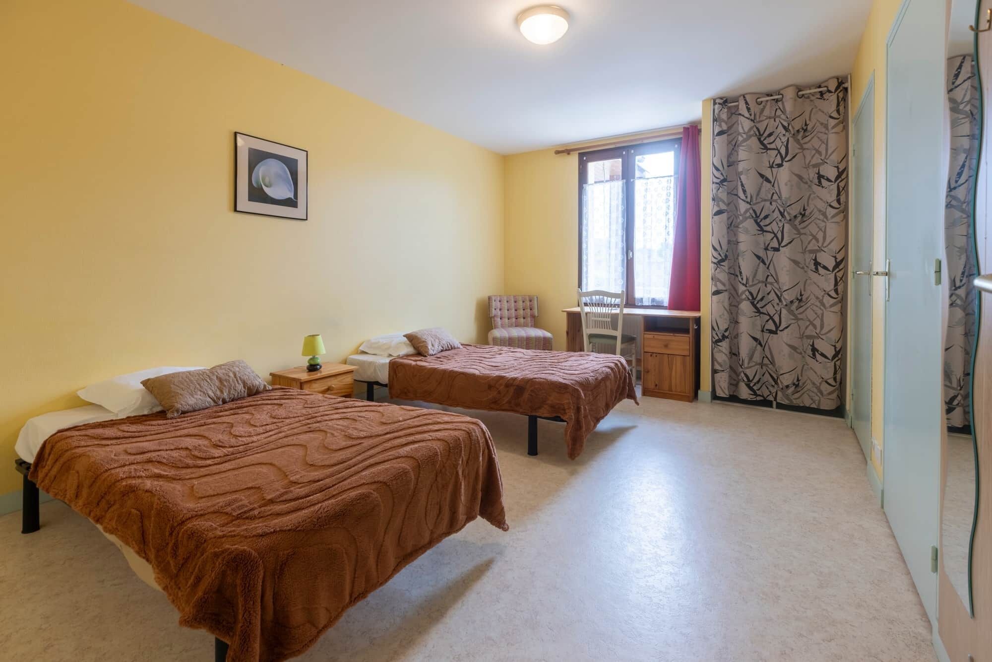 A bedroom with two beds, a desk, a chair, a window with curtains, and a door with a patterned curtain.