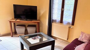A living room with a television on a wooden table, a glass coffee table, a sofa with a cushion, and a window with a view of greenery. - Room 104 - Le Bas Mont Residence (Moulay)