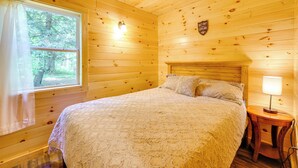 Ein Schlafzimmer einer Holzhütte mit einem Bett, einem Nachttisch, einer Lampe und einem Fenster mit Vorhängen.
