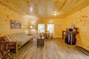A cabin interior with a wooden floor, a sofa, a dining table with chairs, a small kitchenette, and a painting on the wall. - Cabin w/ Lake Access at Letchworth Valley Resort (Bliss)