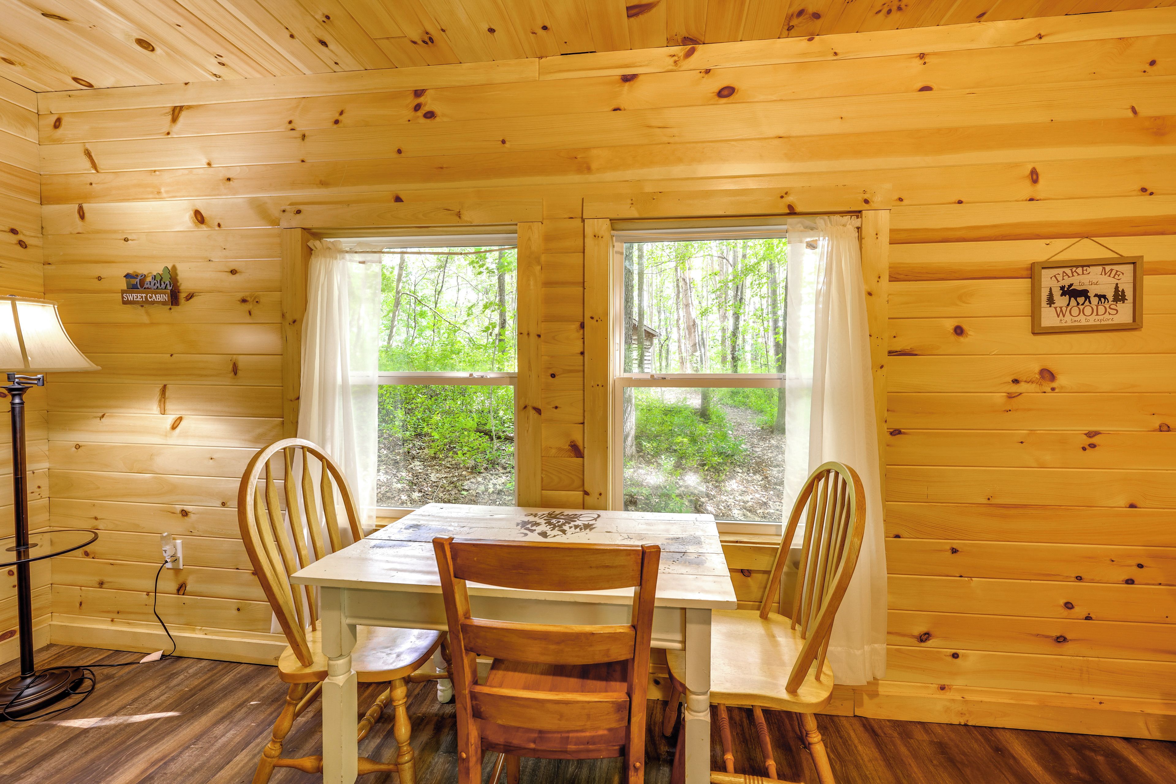 L'intérieur d'un chalet en bois comprend une table à manger, des chaises, une lampe et deux fenêtres donnant sur la forêt.