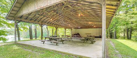 A covered picnic area with picnic tables and benches, surrounded by trees and a lake.