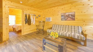 Interior de una cabaña de madera con una cama, un sillón mecedor, un sofá y un cuadro en la pared.