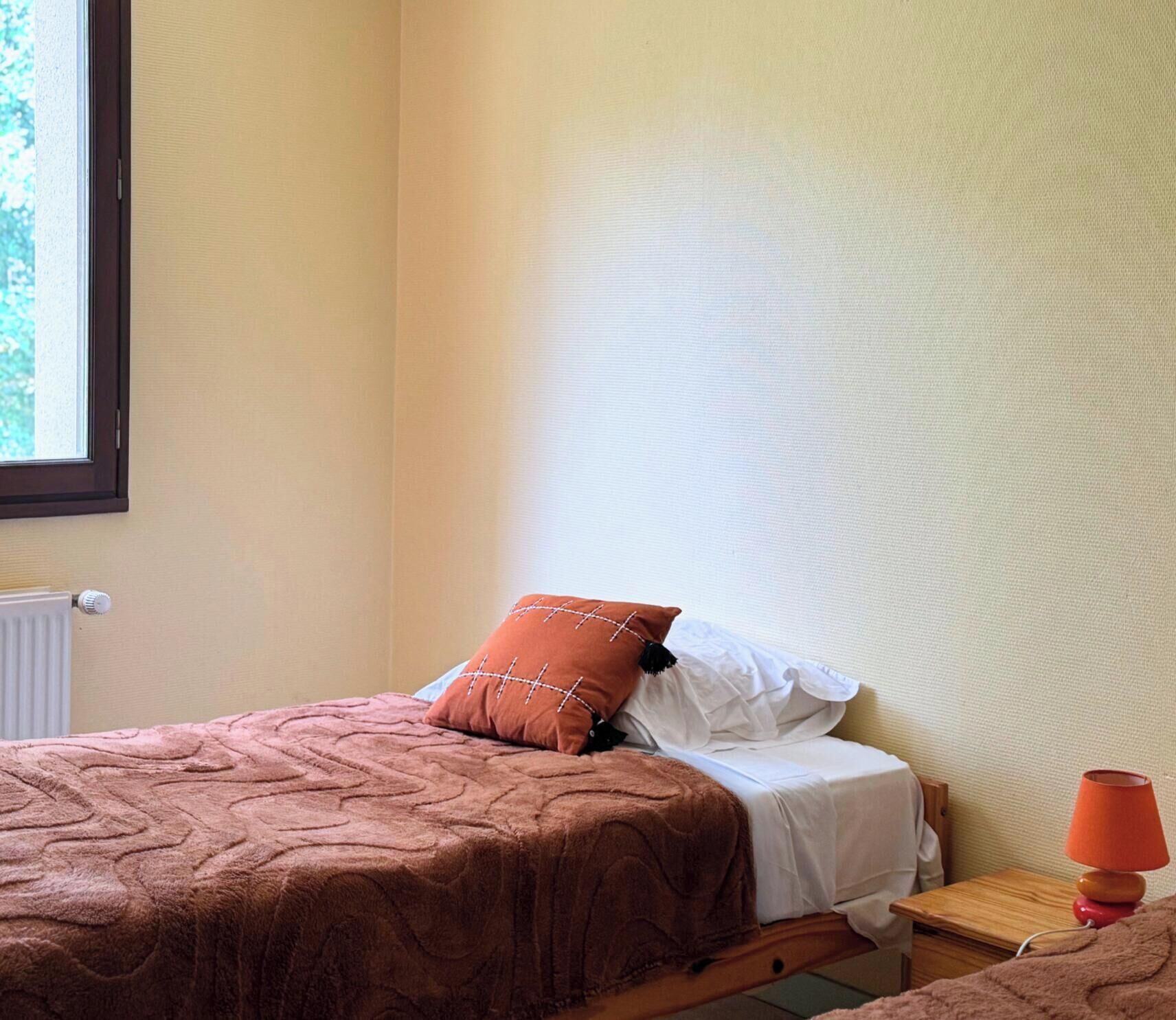 Un letto singolo con una coperta marrone e un cuscino decorativo, un comodino con una lampada rossa e una finestra vista alberi.
