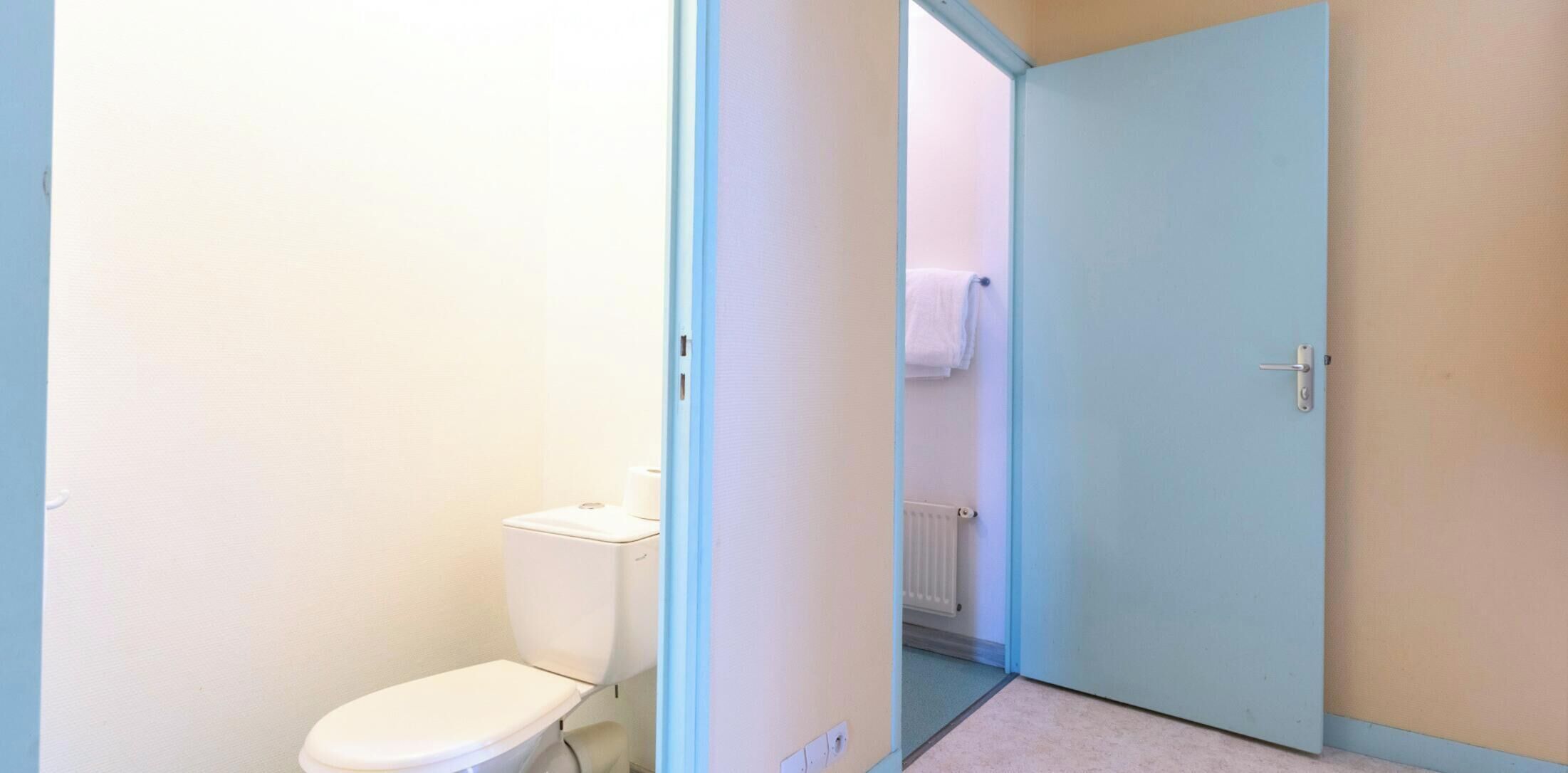 Une petite salle de bain étroite, avec des toilettes, un radiateur et une porte donnant sur une autre pièce.