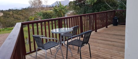 A wooden deck with a glass table and two chairs, overlooking a mountainous landscape.
