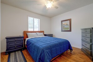 Ein Schlafzimmer mit Bett, Nachttischen, einem Schminktisch, einem Deckenventilator, einem Fenster mit Jalousien und einem gerahmten Bild an der Wand.