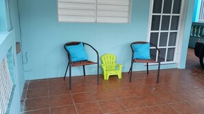Een veranda met twee rieten stoelen en een plastic stoel, een betegelde vloer en een raam met zonwering.