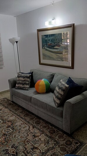 Um sofá cinzento com almofadas estampadas, uma bola de praia colorida, um quadro emoldurado e um abajur.