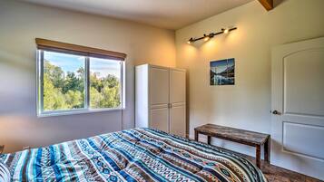 Quarto com uma cama grande, janela com vista para árvores, guarda-roupa, uma mesa pequena e uma porta.