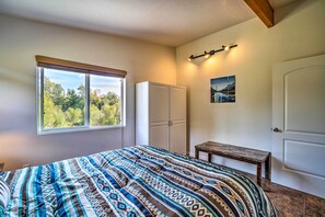 A bedroom with a large bed, a window with a view of trees, a wardrobe, a small table, and a door.