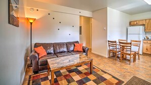 A living room with a brown leather sofa, a coffee table, and a patterned rug. There is a dining area with wooden chairs and a small table. The kitchen is visible with wooden cabinets and a white refrigerator. - Steps to Fritz Creek! 2-Unit Family Retreat (Fritz Creek)
