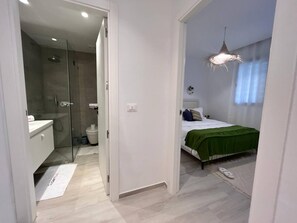 Ein modernes Schlafzimmer mit Bett, Nachttisch, Pendelleuchte und Blick auf ein Badezimmer mit Dusche und Toilette.