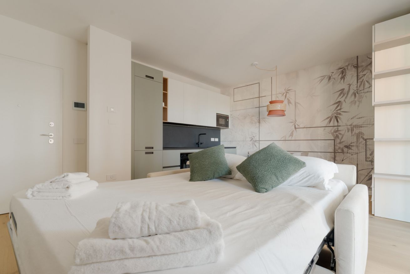 A hotel room with a bed, pillows, a bedside table, a wardrobe, and a view of the kitchen.