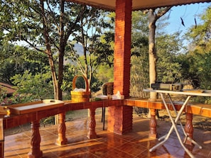 An outdoor patio with a wooden table and chairs, surrounded by trees and a brick pillar.