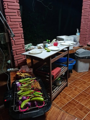 Una cocina con una parrilla, una mesa con platos y aderezos, y un cubo de basura.