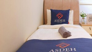 Una camera d'albergo con un letto, un cuscino con il logo ASDEH SUTES e un asciugamano piegato con cura.