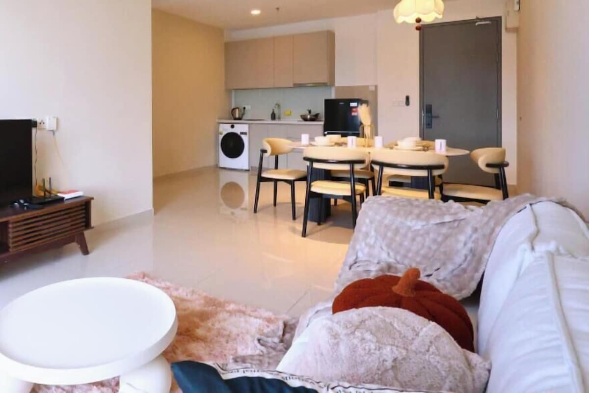 Ein modernes Wohnzimmer mit einer Couch, einem Couchtisch und einem Essbereich mit Stühlen. Eine Deckenlampe, eine Klimaanlage und eine Küche sind im Hintergrund zu sehen.