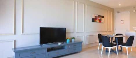 Uma sala moderna com televisão de tela plana em um móvel azul, área de jantar com mesa redonda e cadeiras, e uma porta ao fundo.
