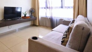 A living room with a flat-screen TV, a sofa, a fan, and a window with curtains. - I-City Shah Alam Residences By Asdeh (Shah Alam)
