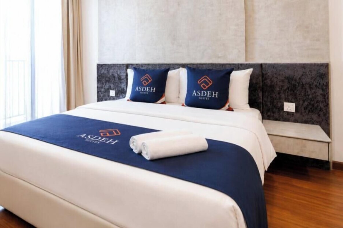 Ein Hotelzimmer mit einem großen Bett, zwei Kissen mit dem Logo des Hotels, einer ordentlich bezogenen Bettdecke und einem Nachttisch mit einer Lampe.
