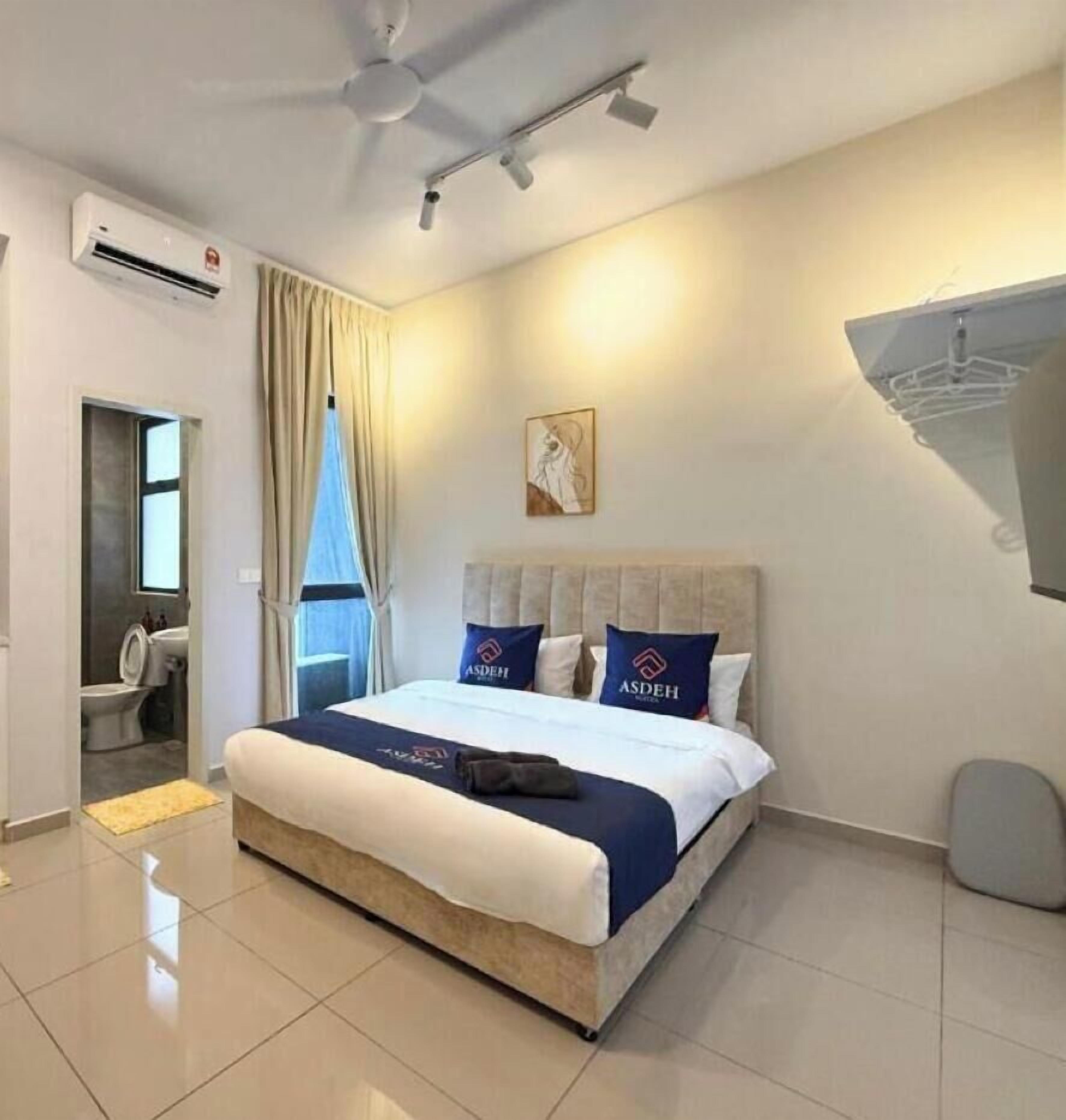 Habitación de hotel con cama, ventilador de techo, equipo de aire acondicionado, ventana con cortinas y lámpara fijada a la pared.