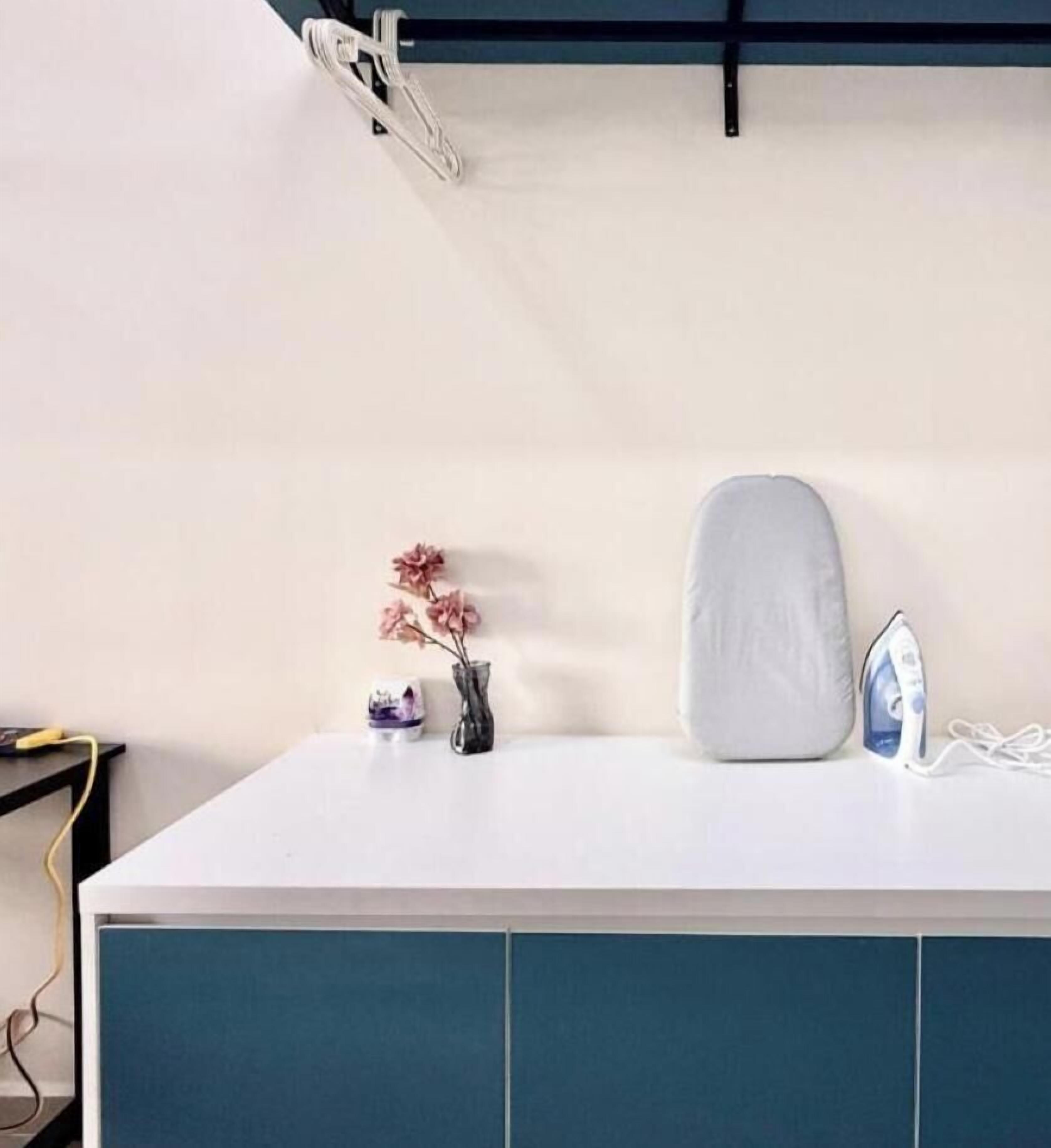 A white countertop with a blue cabinet below, a white cushion, a vase with pink flowers, and an iron on the right side.