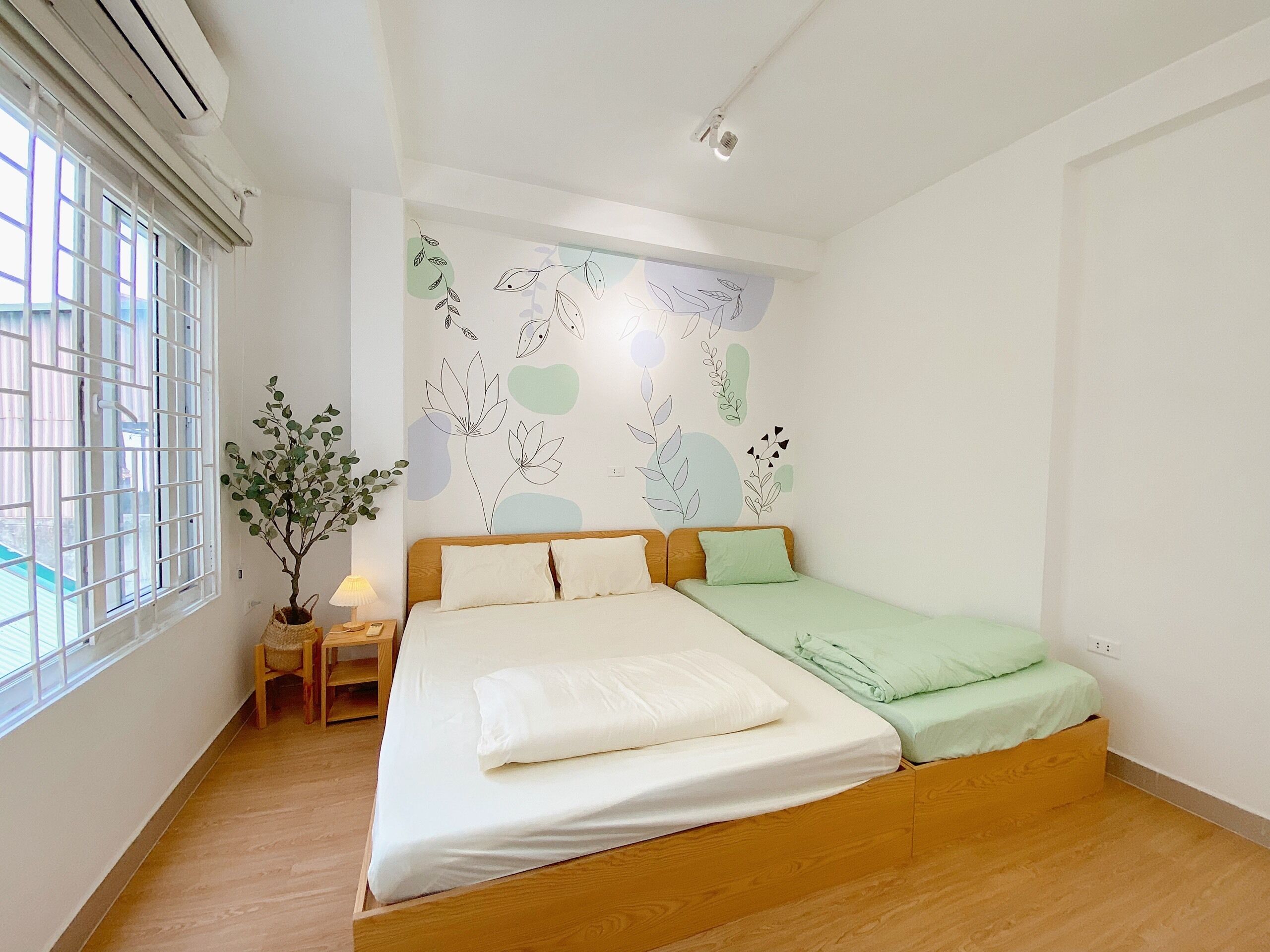 Un dormitorio con cama de madera, una mesita de noche, una lámpara, una planta en maceta y una ventana con barrotes.