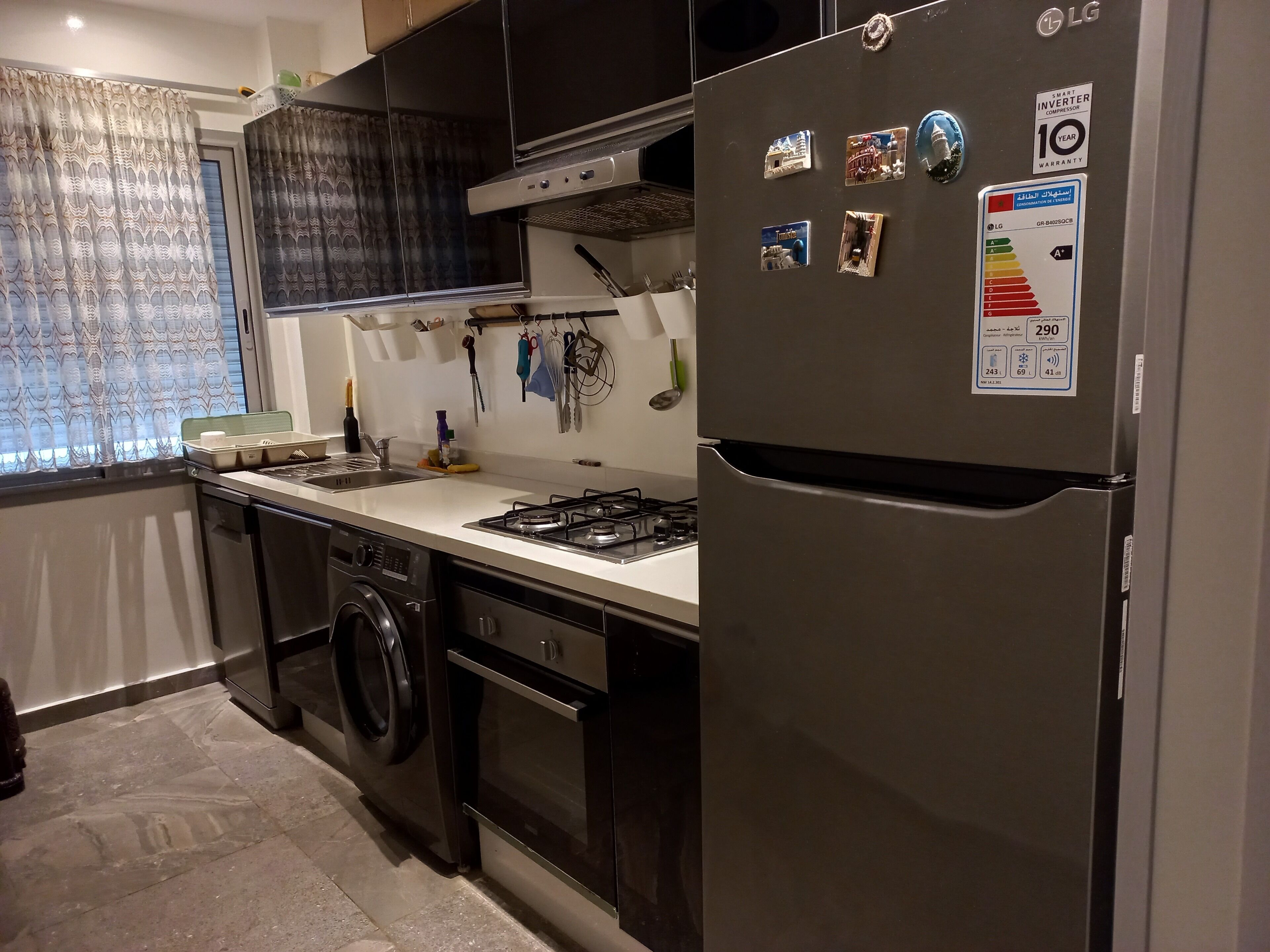 A compact kitchen with a stainless steel refrigerator, a gas stove, a built-in oven, and a washing machine.