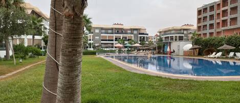 A swimming pool with lounge chairs and umbrellas, surrounded by greenery and multi-story buildings.