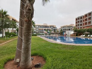 Ein Schwimmbecken mit Liegestühlen und Sonnenschirmen, umgeben von Grünflächen und mehrstöckigen Gebäuden.