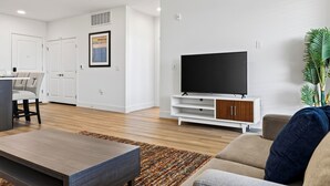 A modern living room with a flat-screen TV, a coffee table, and a sofa. There is a dining area with chairs and a table, and a kitchen area with cabinets and a countertop.