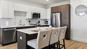 Una cocina moderna con armarios blancos, un refrigerador de acero inoxidable, un microondas y una cafetera. Cuenta con una zona de comedor con una isla blanca y dos sillas.