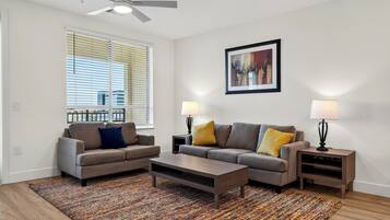 A modern living room with a sofa set, a coffee table, two side tables with lamps, a ceiling fan, and a large window with blinds.