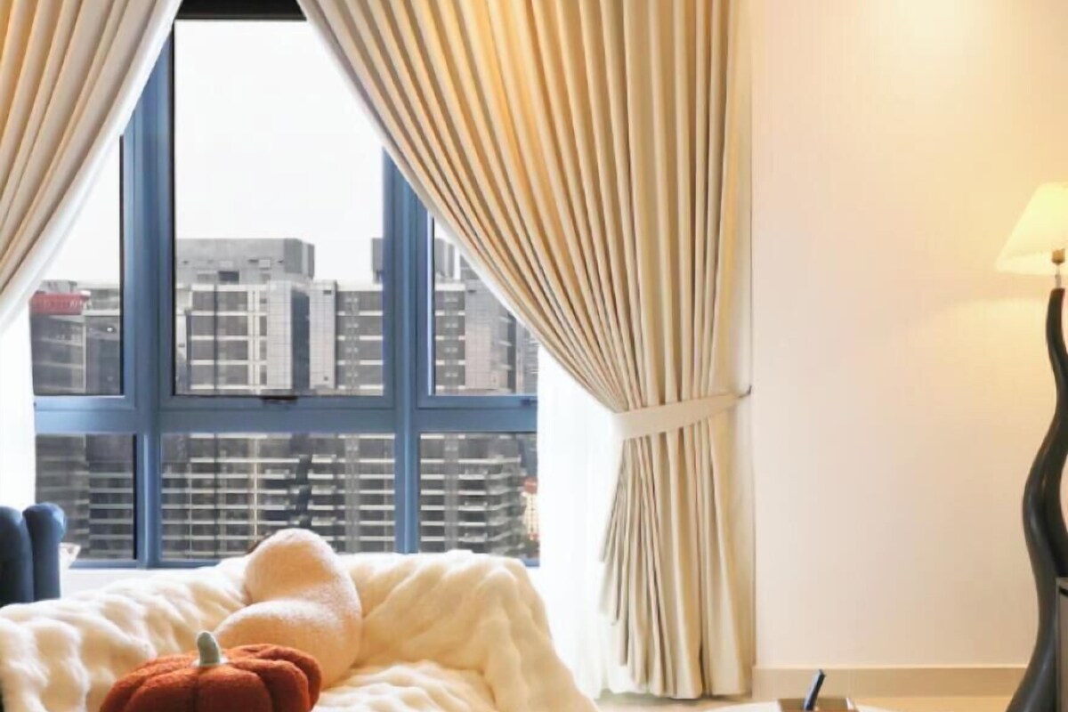 A hotel room with a bed, a white table, and a view of buildings through large windows.