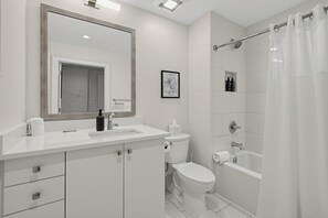 A modern bathroom with a white sink, mirror, toilet, and shower. - Historic Saddlery Condo • 2 Mins to Broadway + Near Printer's Alley | Key Note (Nashville)