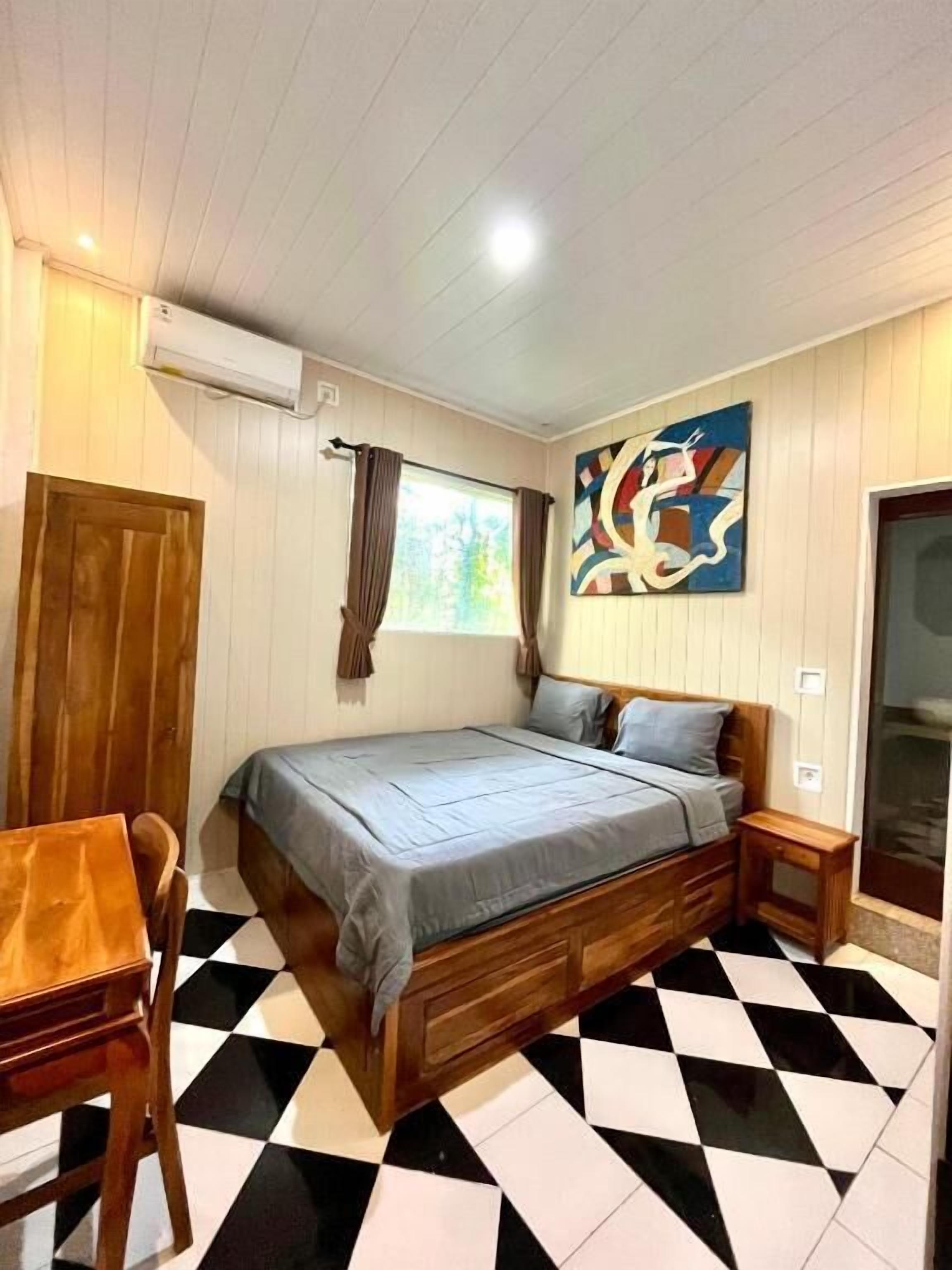 Una habitación con cama, una silla de madera, una mesita, una ventana con cortinas y un aire acondicionado de pared.