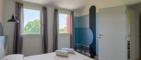 A bedroom with a large bed, two windows with curtains, a bedside lamp, and a door leading to another room.